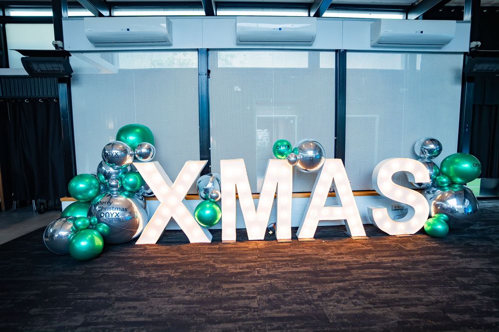 Large illuminated XMAS letters decorated with green and silver balloon garlands on a dark floor against a blue backdrop.