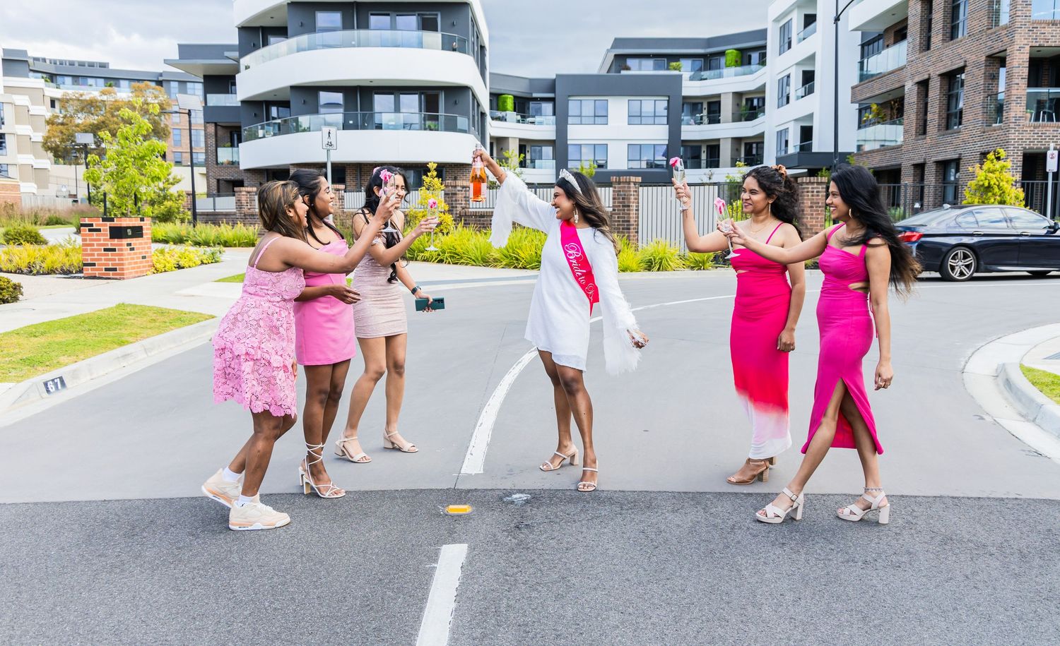 A group in pink dresses playfully poses together on a residential street with modern apartment buildings in the background.
