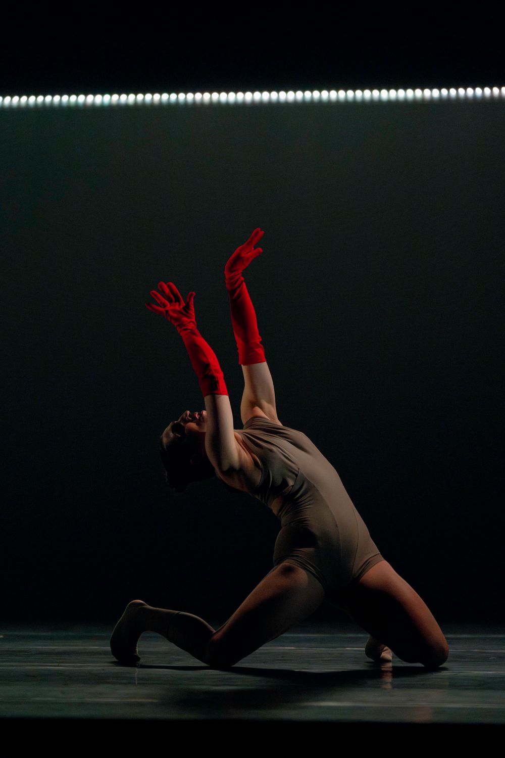 A dancer in red gloves performs an expressive backbend under a line of bright stage lights.