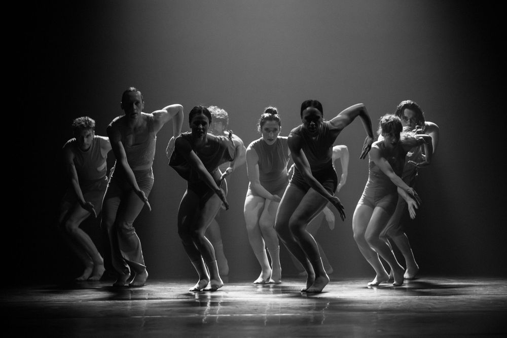 Dancers perform an energetic contemporary dance routine on a darkened stage illuminated by dramatic spotlighting.