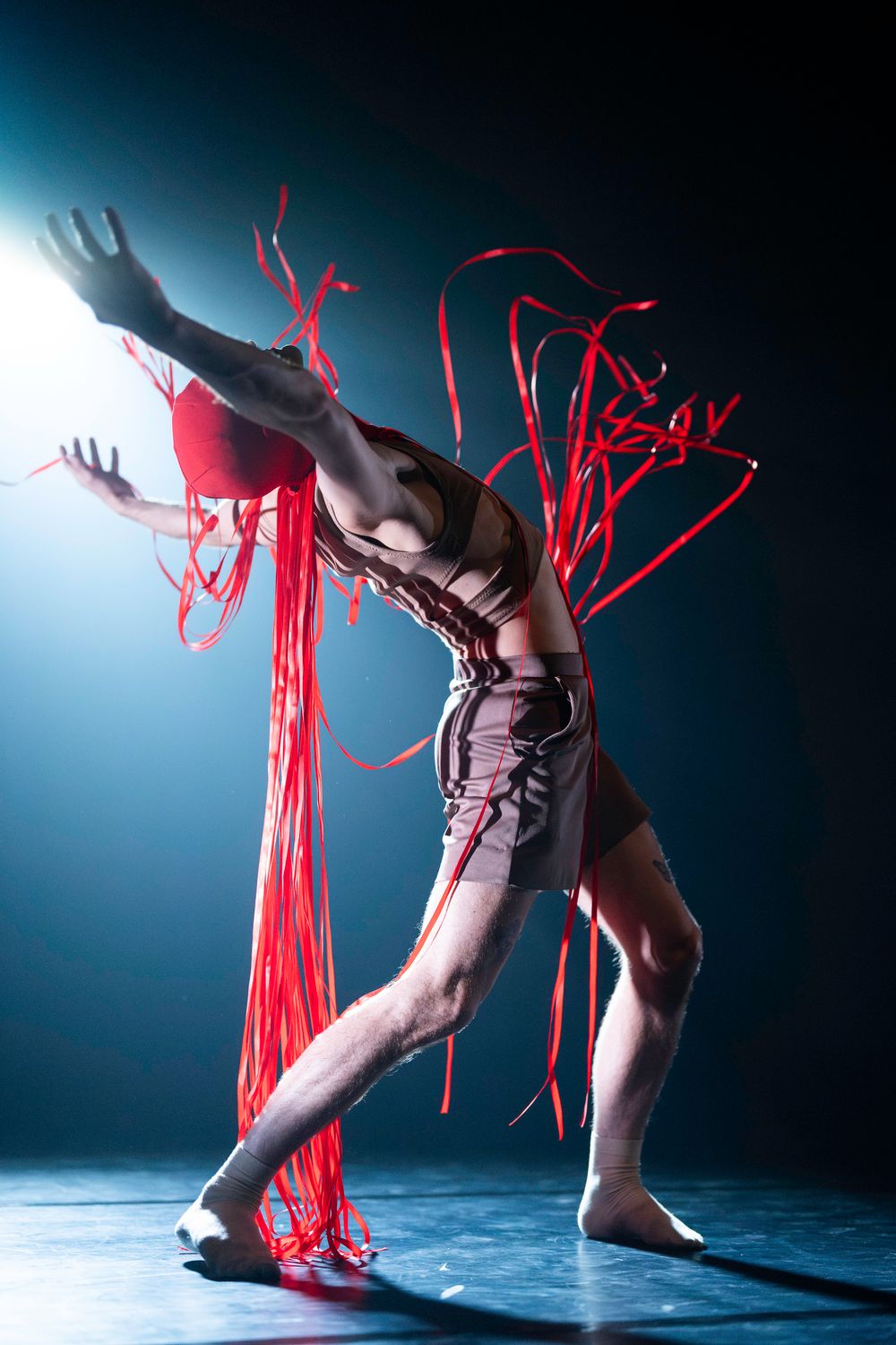 Stylistic contemporary dancer in dramatic pose with flowing red ribbons against moody blue studio lighting.