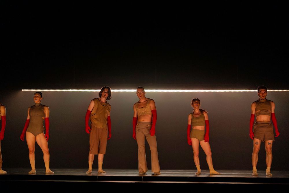 Line of dancers in red costumes performing on stage with minimalist lighting design.
