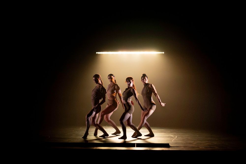 Dancers moving in sequence under dramatic spotlight on dark stage.