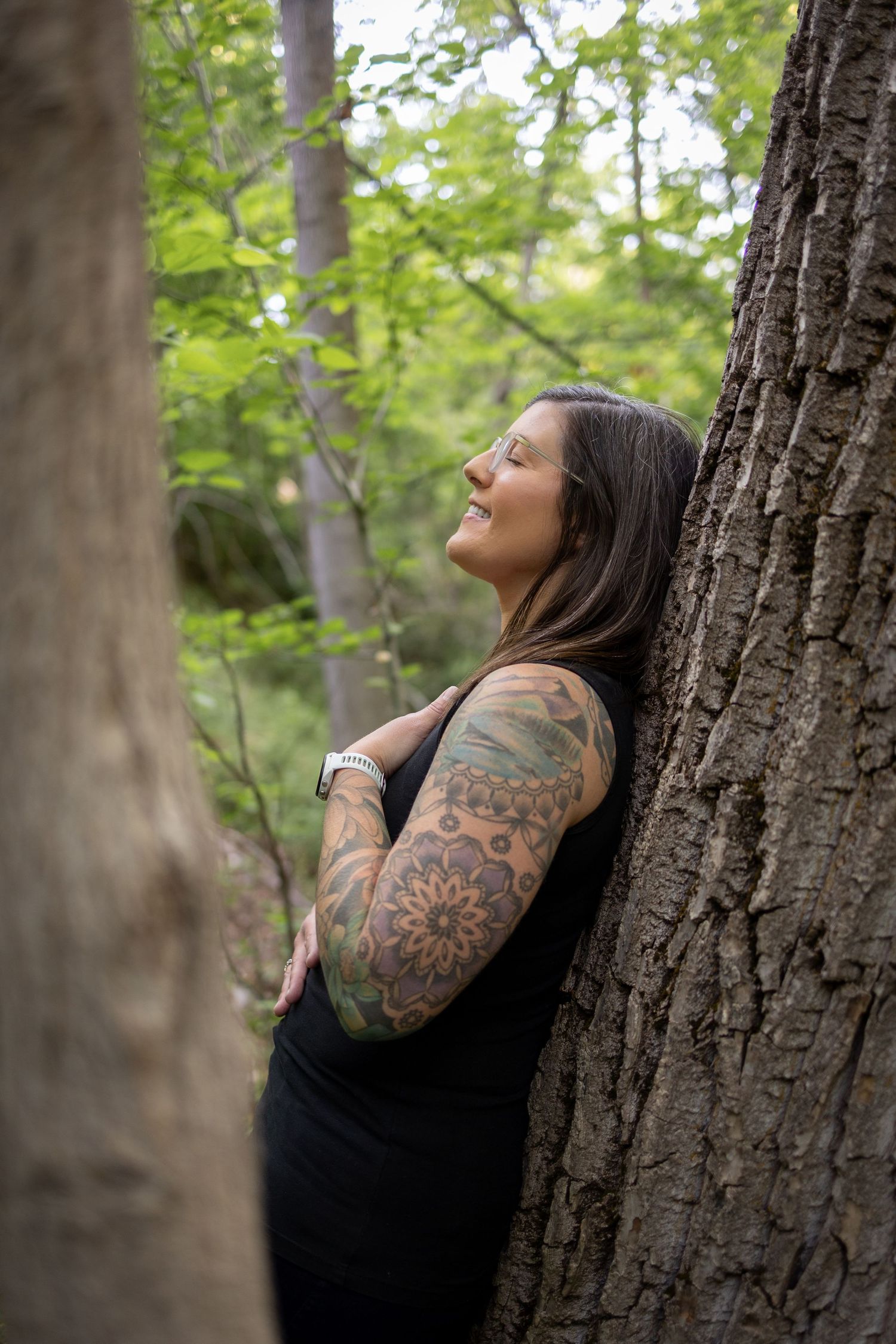 Person with colorful tattoo sleeve leans against tree trunk in peaceful forest setting during summer.