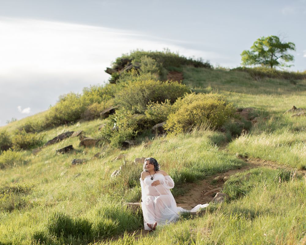 A figure in a flowing white dress sits peacefully on a green hillside beneath cloudy skies.