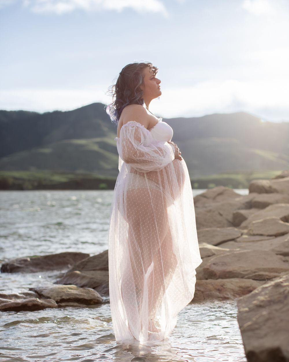 Ethereal figure in flowing white dress poses gracefully on rocky coastline at sunset creating dreamy silhouette.