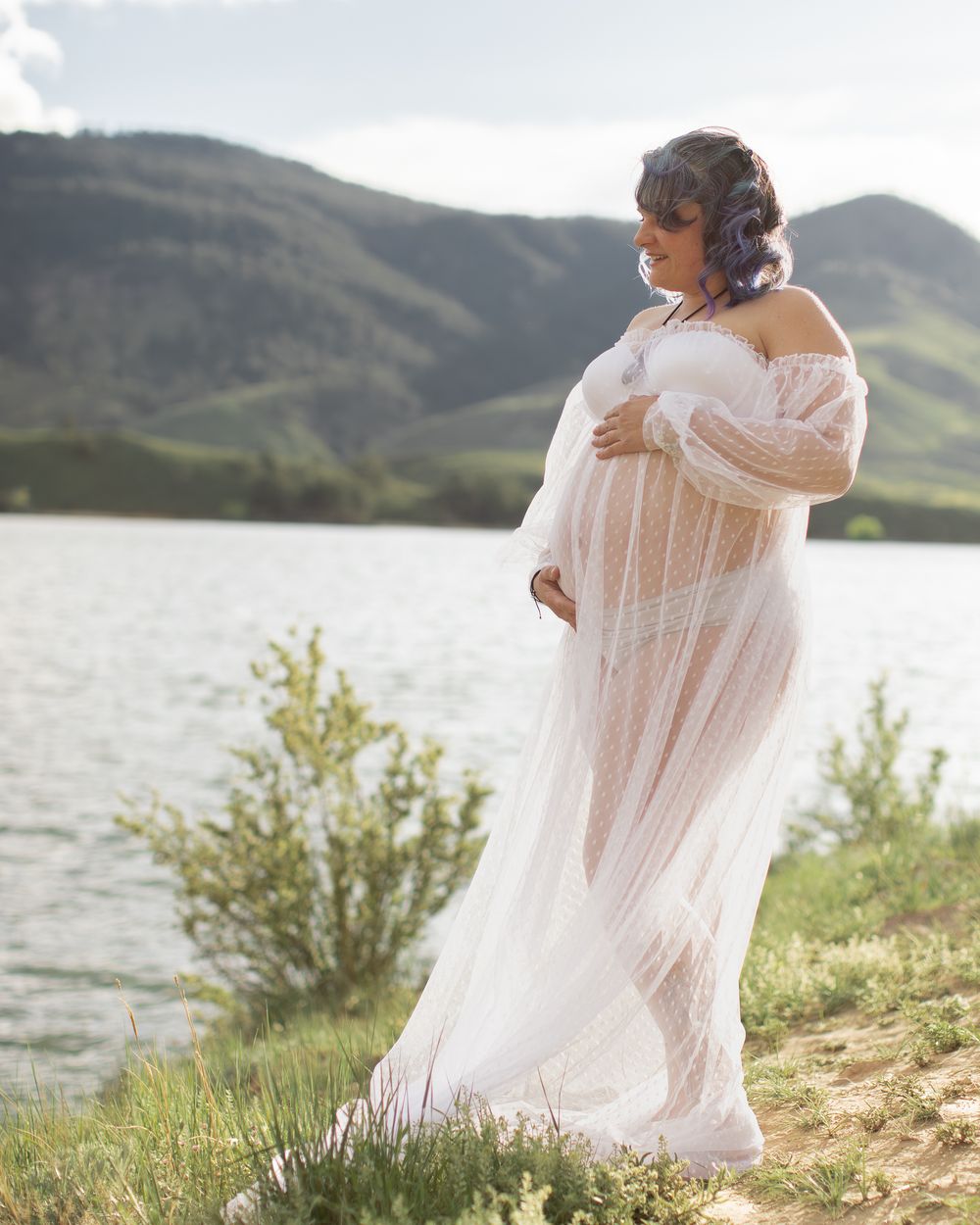 Ethereal white maternity dress flows in the breeze as expectant mother stands by lakeside with mountains in background.