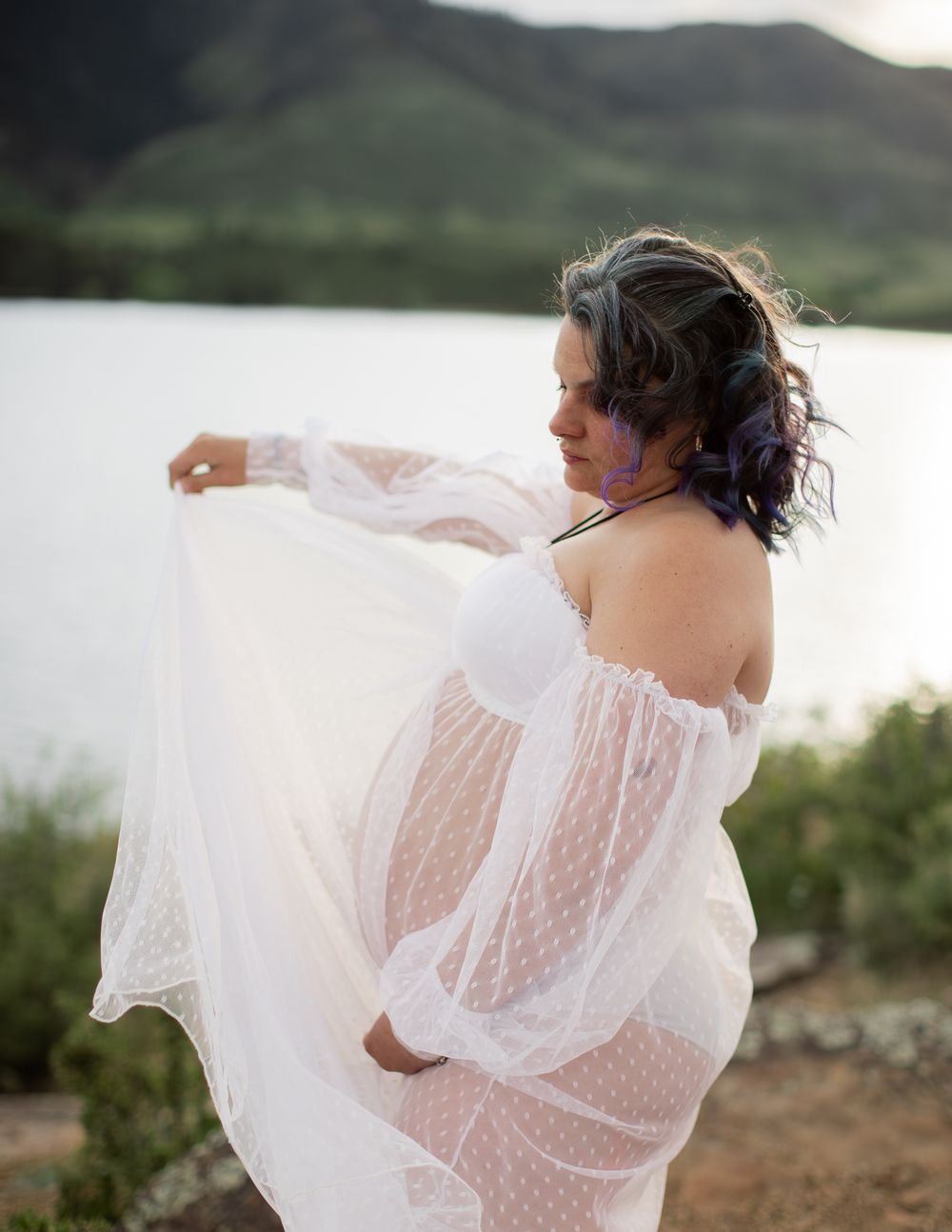 Figure in a sheer white lace dress poses gracefully by a lakeside with mountains in the background.