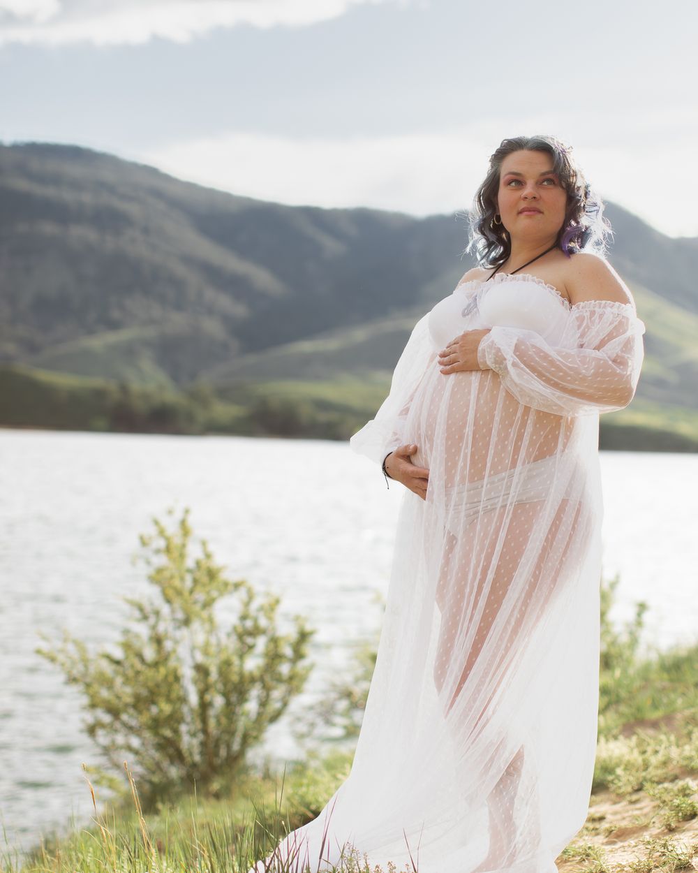 Person wearing flowing white dress poses elegantly by lake with mountains in background on sunny day.