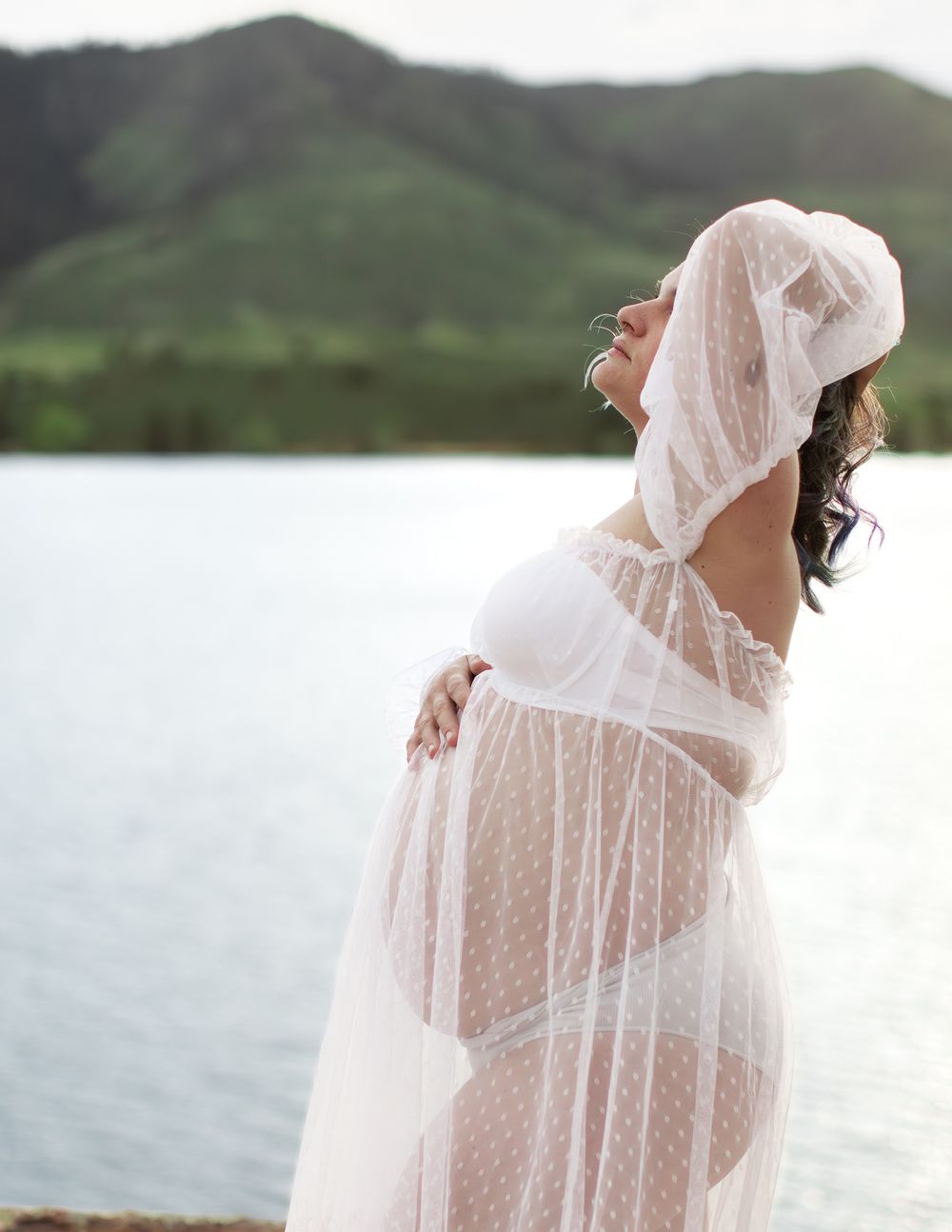 A person in a flowing white sheer dress and veil stands gracefully by a scenic lake with mountains in the background.