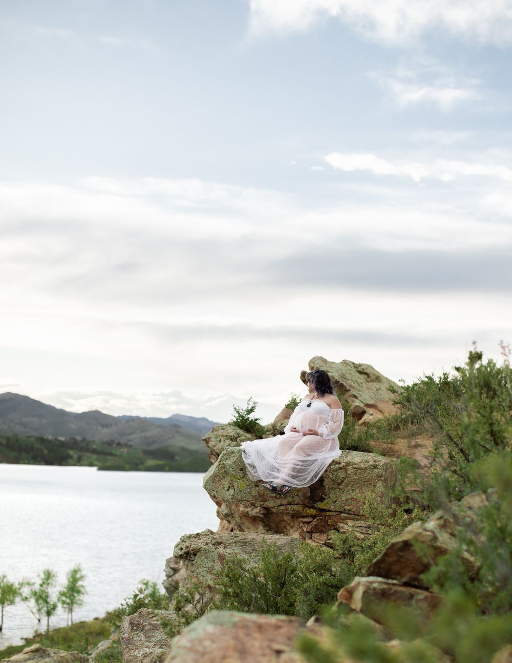 A figure in a white dress sits peacefully on a rocky outcrop overlooking a scenic mountain lake on a cloudy day.