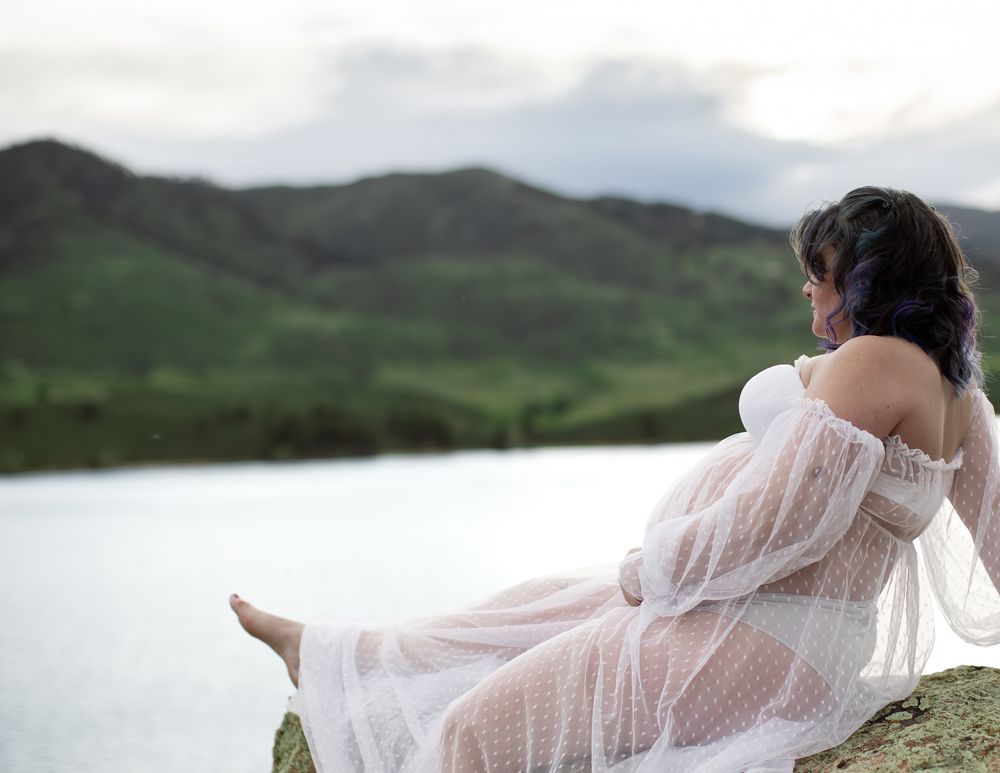 A person in a flowing white dress sits near a serene lake surrounded by rolling green mountains.