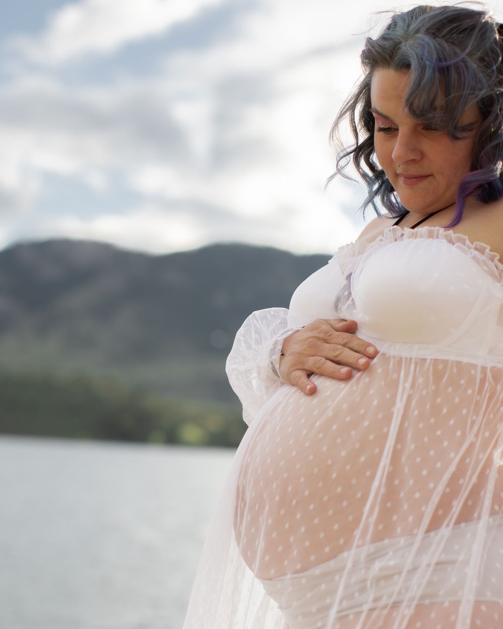 Expectant mother in sheer white lace gown poses near a lake with mountains in the background.