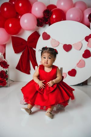 Toddler girl in red dress sits among Valentine's Day balloons and heart decorations.