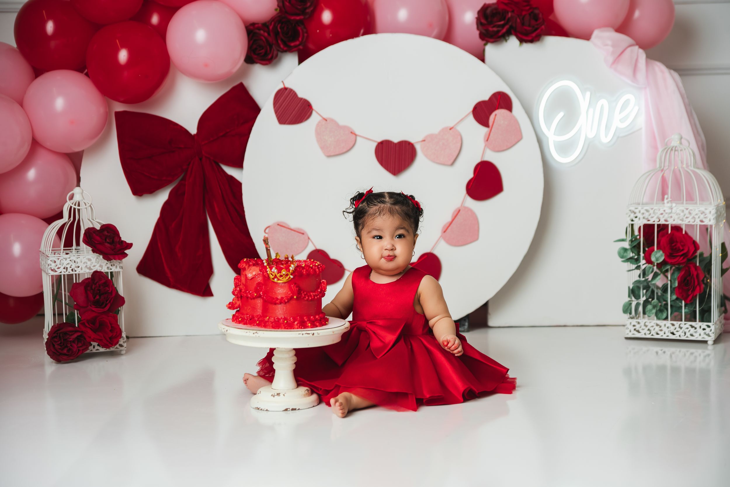 Baby girl in red dress sits by a red smash cake at a Valentine's Day first birthday party with balloon decorations.