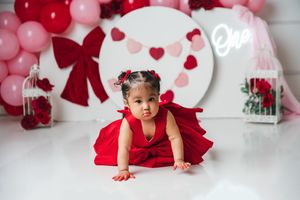 Baby girl in red dress crawls on white floor with Valentine heart balloons and decorations for first birthday photo.