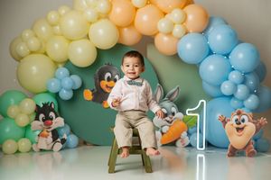 Baby boy sits on chair at 1st birthday with Looney Tunes plush toys and colorful balloon arch.