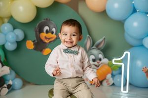 Smiling baby boy in bow tie celebrates first birthday with balloon arch, cartoon characters, and glowing number one.