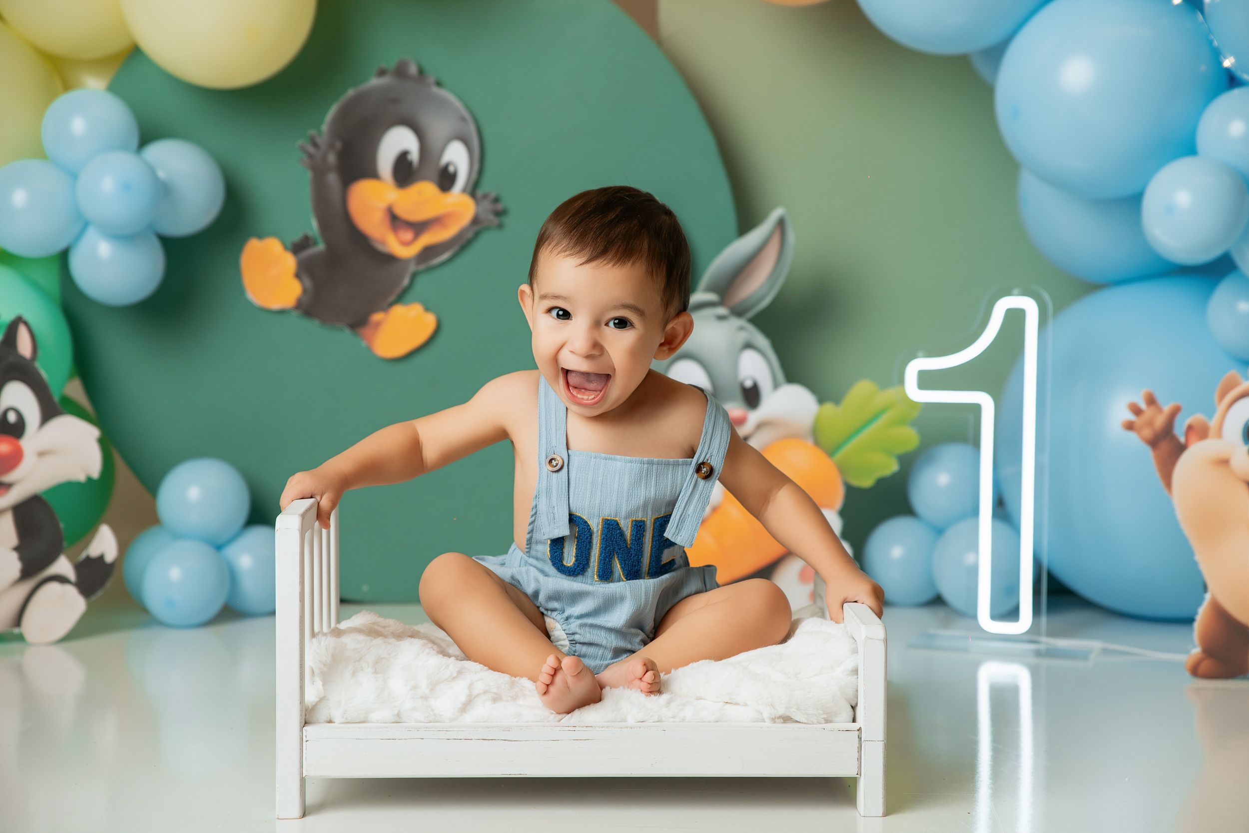 Baby boy in blue overalls sits on white mini bed at his first birthday cake smash photo session with balloon backdrop.