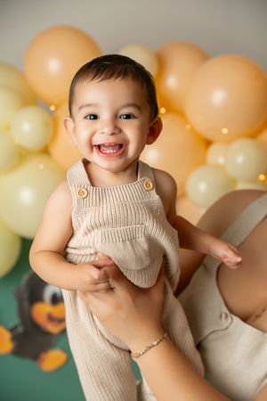 Smiling baby in beige knit overalls held up by mother with yellow balloons in background.