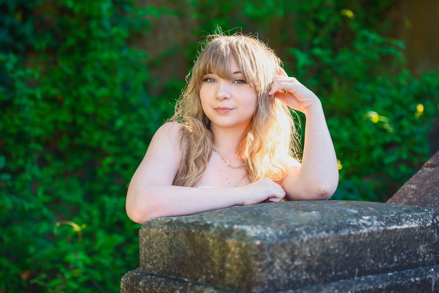Young woman with blonde hair leans against stone wall in outdoor garden setting with green foliage background.