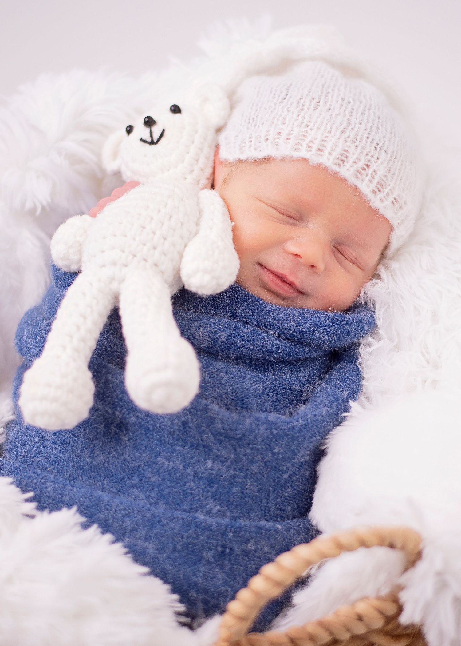 Newborn sleeping peacefully with white knit hat and plush bear toy on blue blanket surrounded by soft white material.