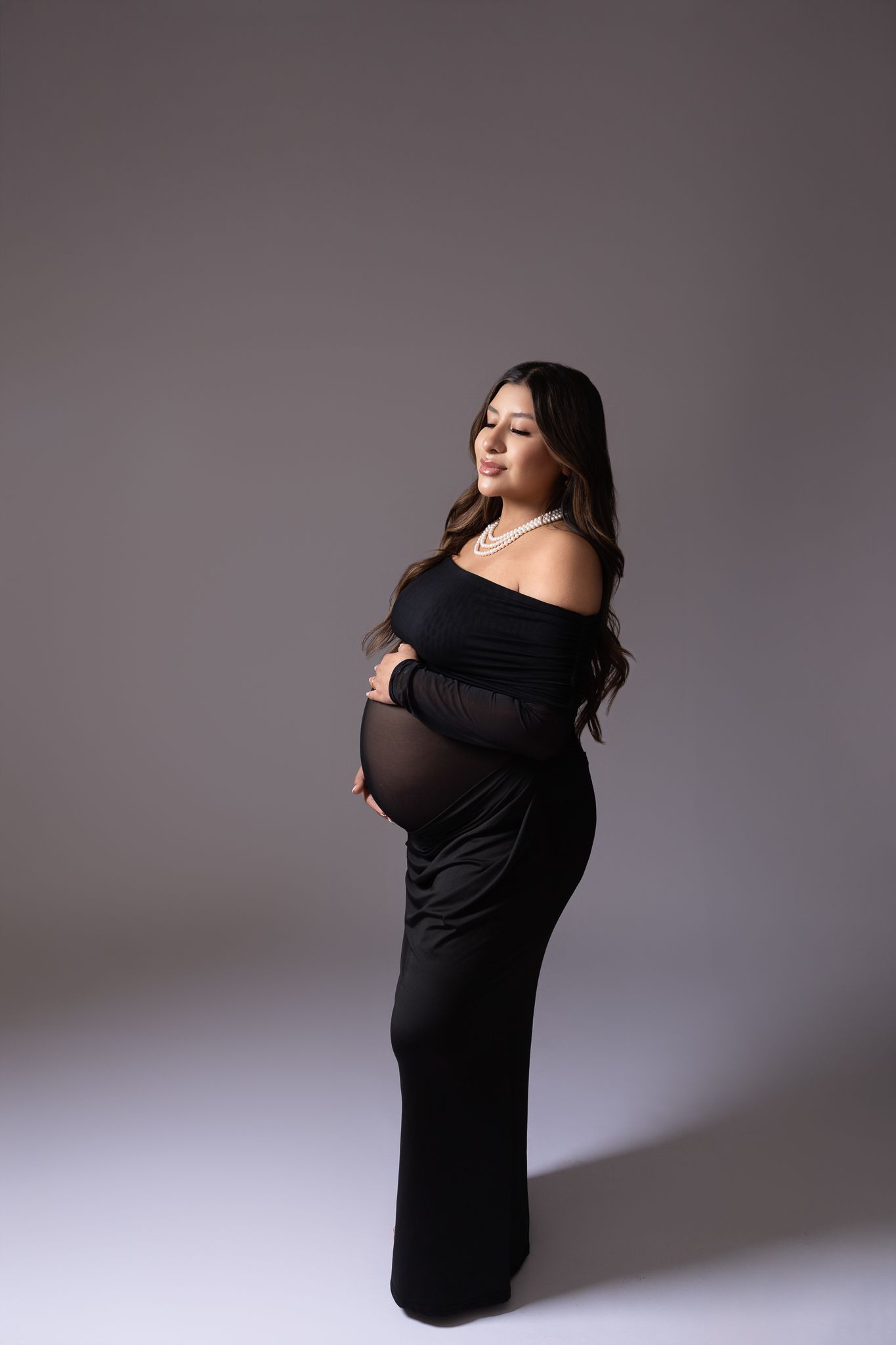 Dramatic maternity portrait of woman in black off-shoulder gown against dark background.
