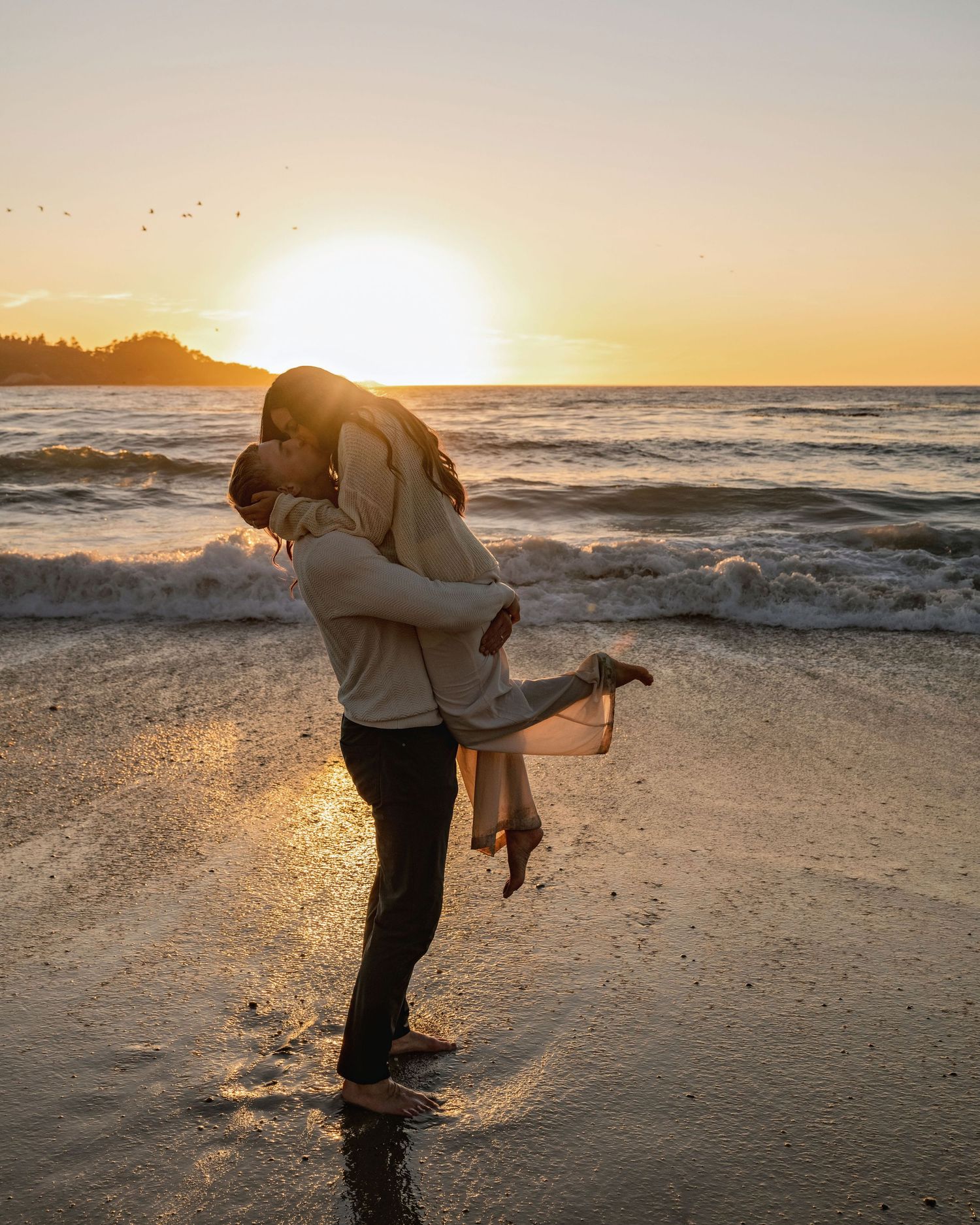 Couple shares a romantic moment on the beach at sunset with dramatic ocean waves in background.