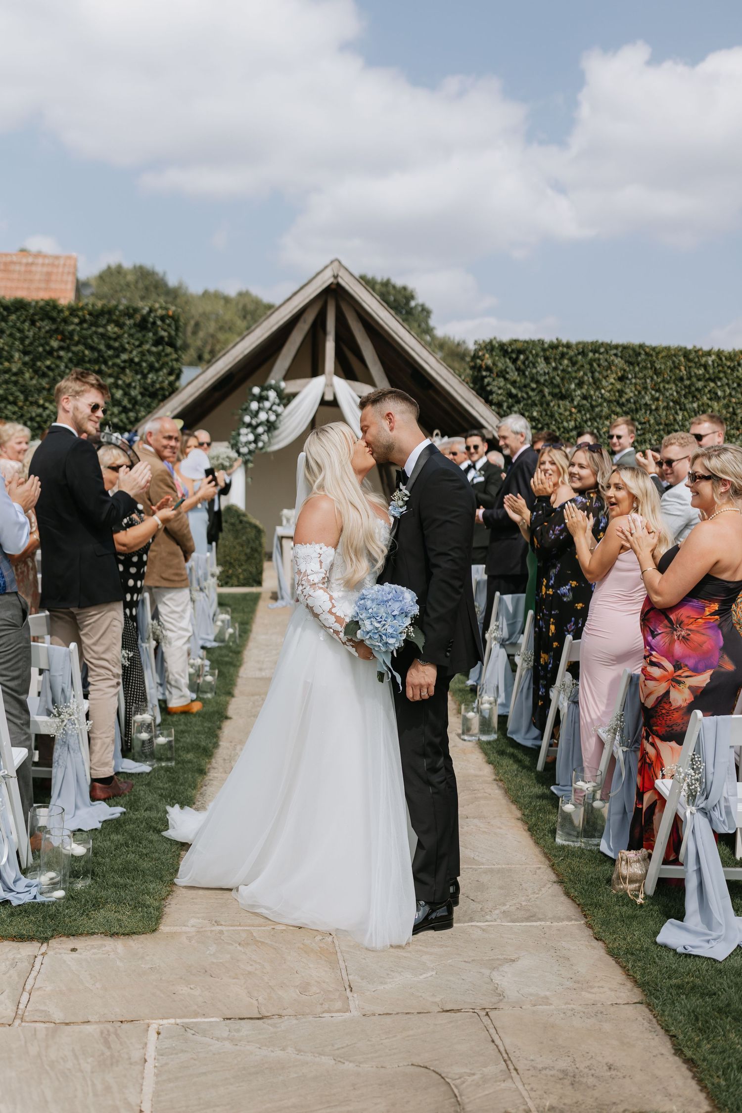 Wedding guests line both sides of an outdoor pathway cheering as newlyweds walk between them during a daytime celebration.