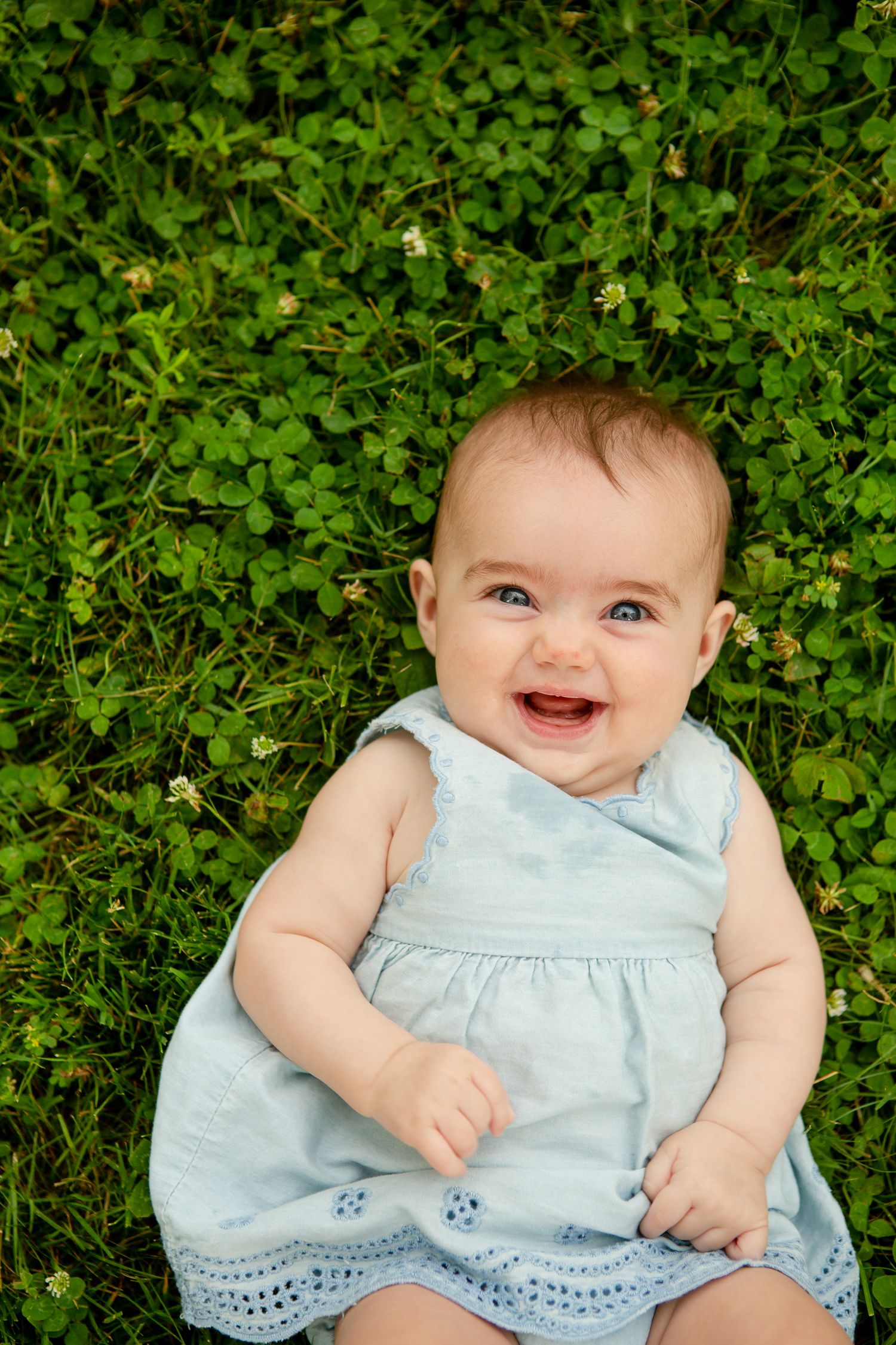 Baby in light blue dress smiles while lying on green clover ground.