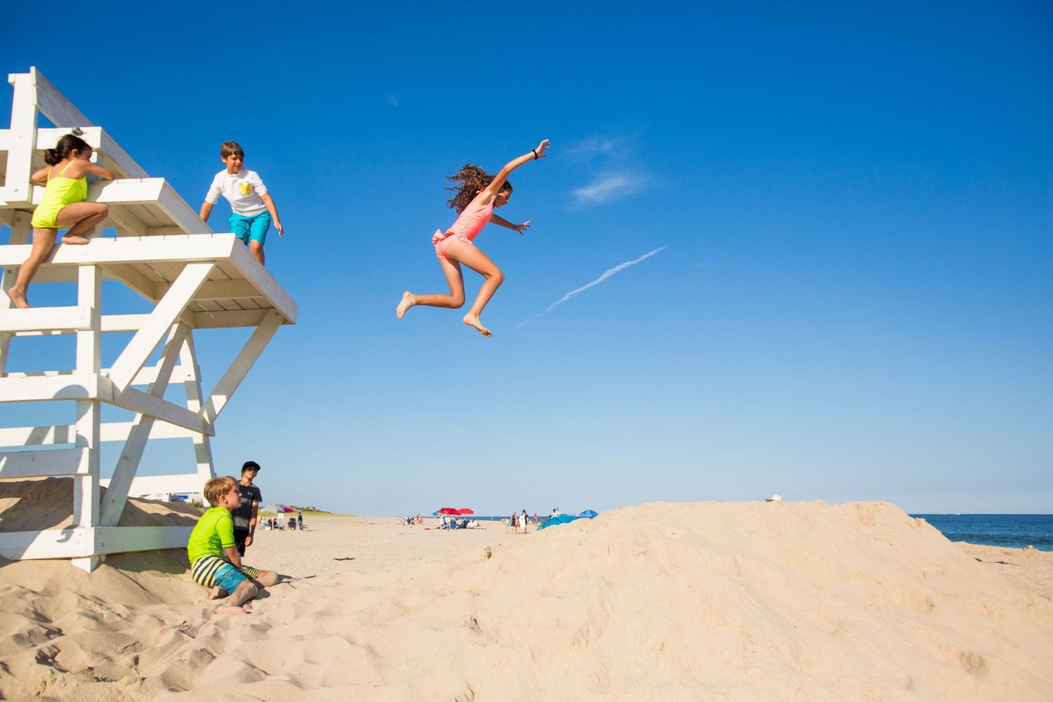 People jumping from a lifeguard tower into sand on a sunny beach day.