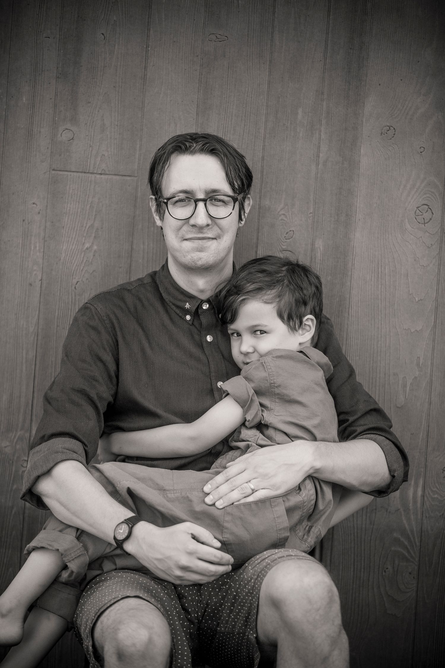 Black and white portrait of parent and child sitting together against wooden wall.