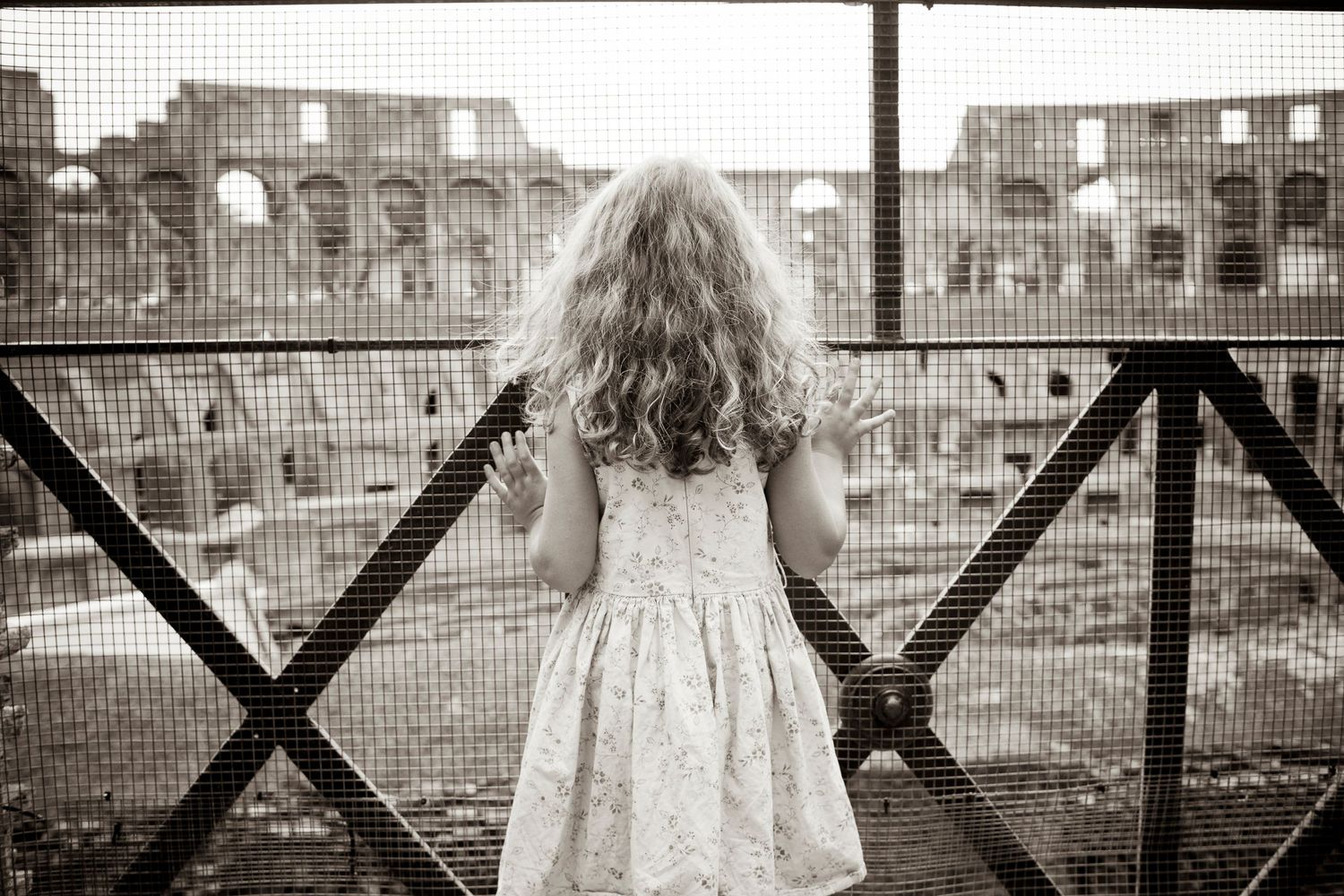 A figure in a dress stands at railing overlooking old brick buildings in black and white photograph.