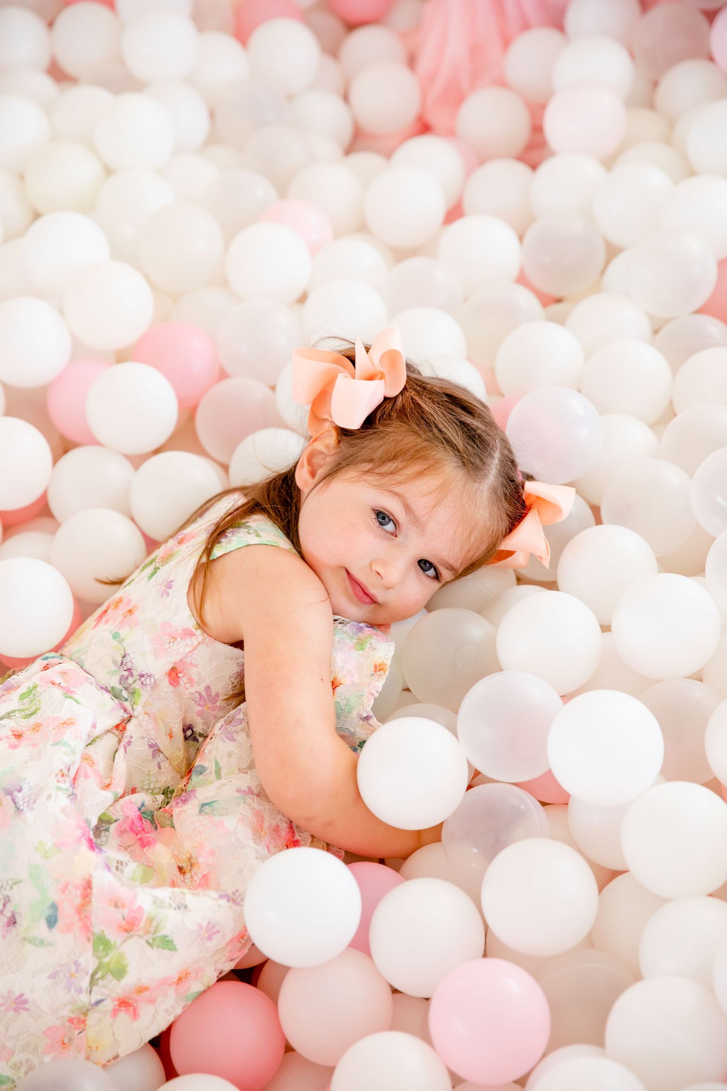 A child in floral dress lies among pink and white ball pit balls.