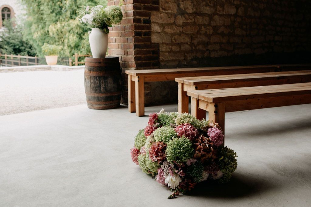 Floral arrangement of hydrangeas on floor beside wooden benches and barrel at outdoor venue.