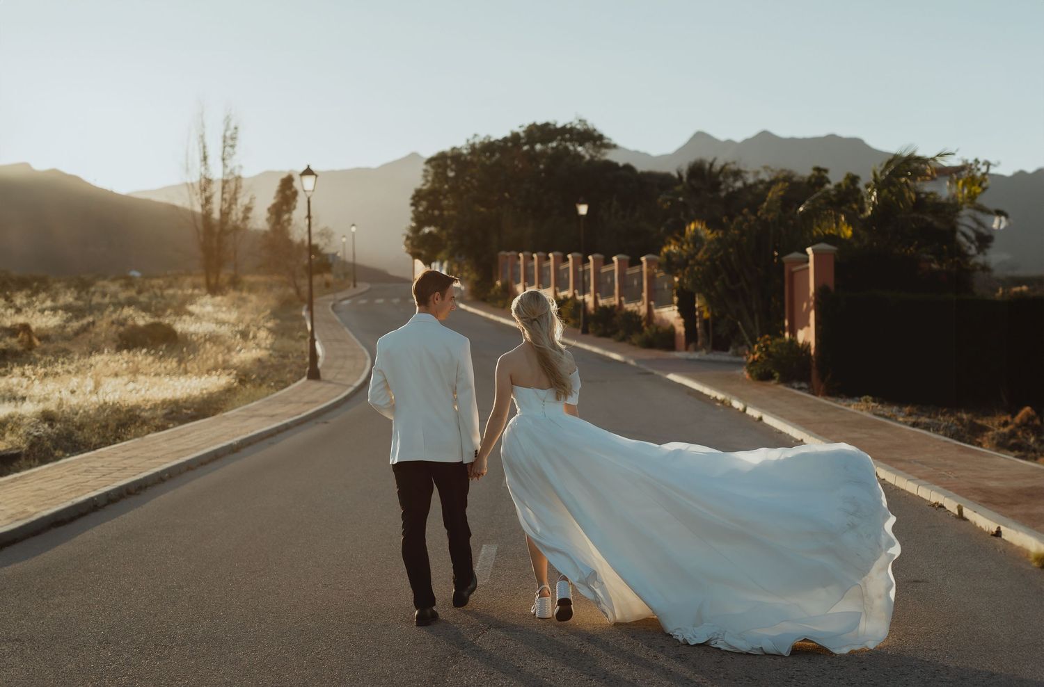 A couple in wedding attire walks together on a winding desert road during golden hour.