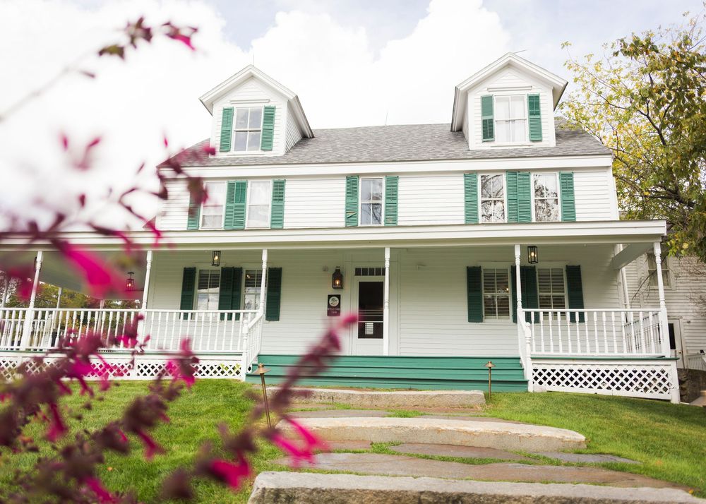 Long white farmhouse with green shutters and wraparound porch in countryside setting.
