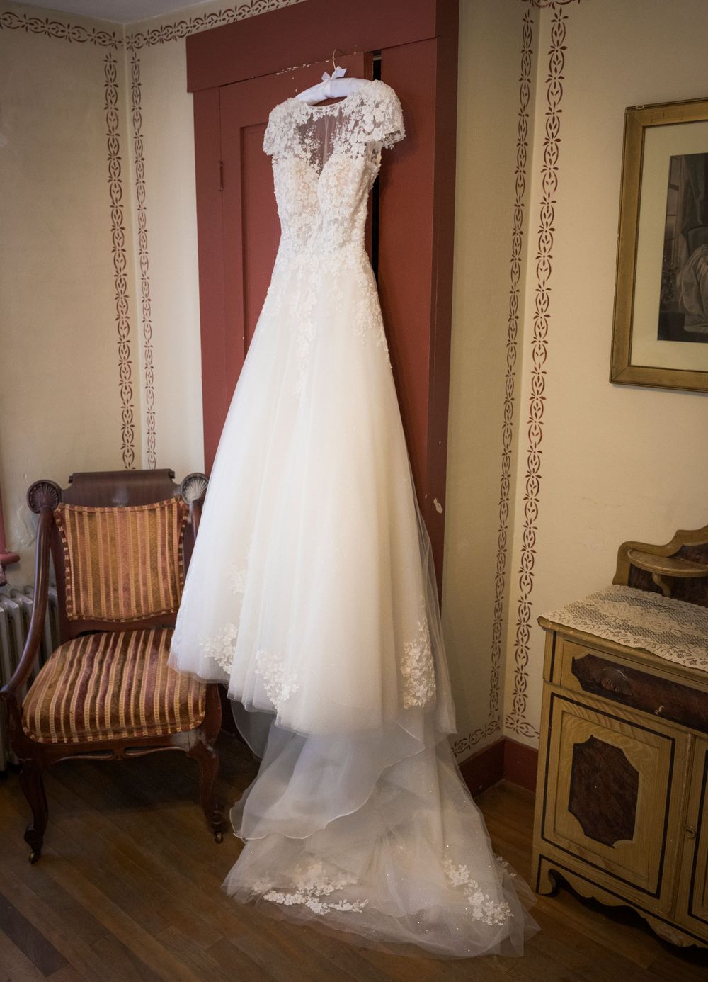 An elegant lace wedding dress with cap sleeves hangs on display in a vintage-styled room.