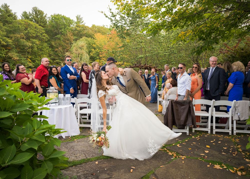 Wedding ceremony guests seated outdoors on white chairs along a tree-lined path in autumn.