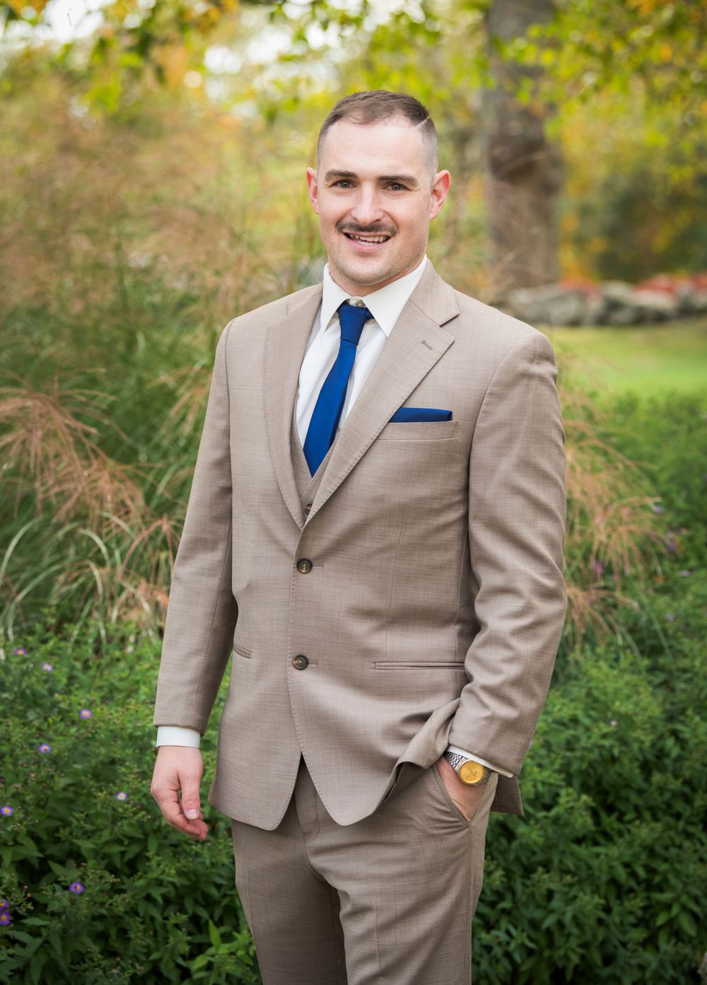 Groom in tan suit with blue tie poses in garden setting during autumn wedding photoshoot.