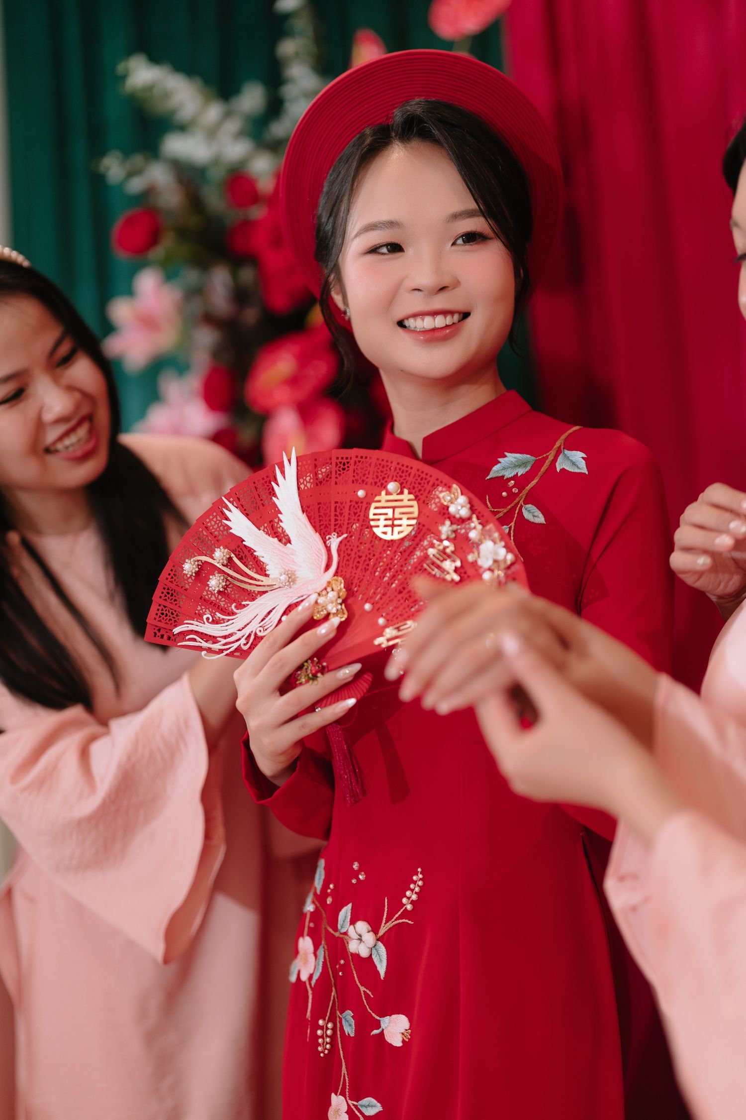Smiling Vietnamese bride in red ao dai receives decorative red fan at traditional wedding ceremony.