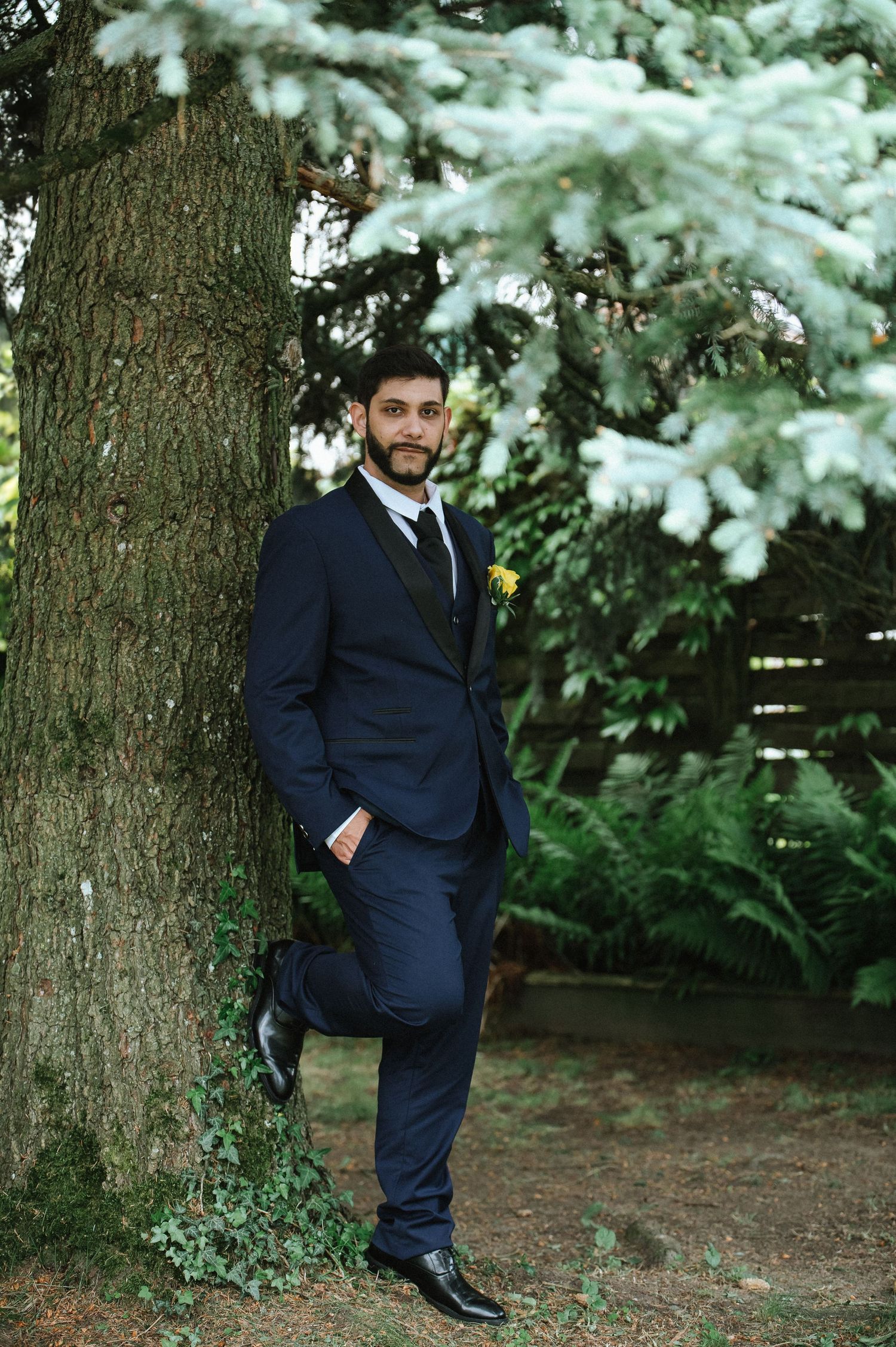 A groom in a navy suit poses against a large tree trunk with evergreen branches overhead.