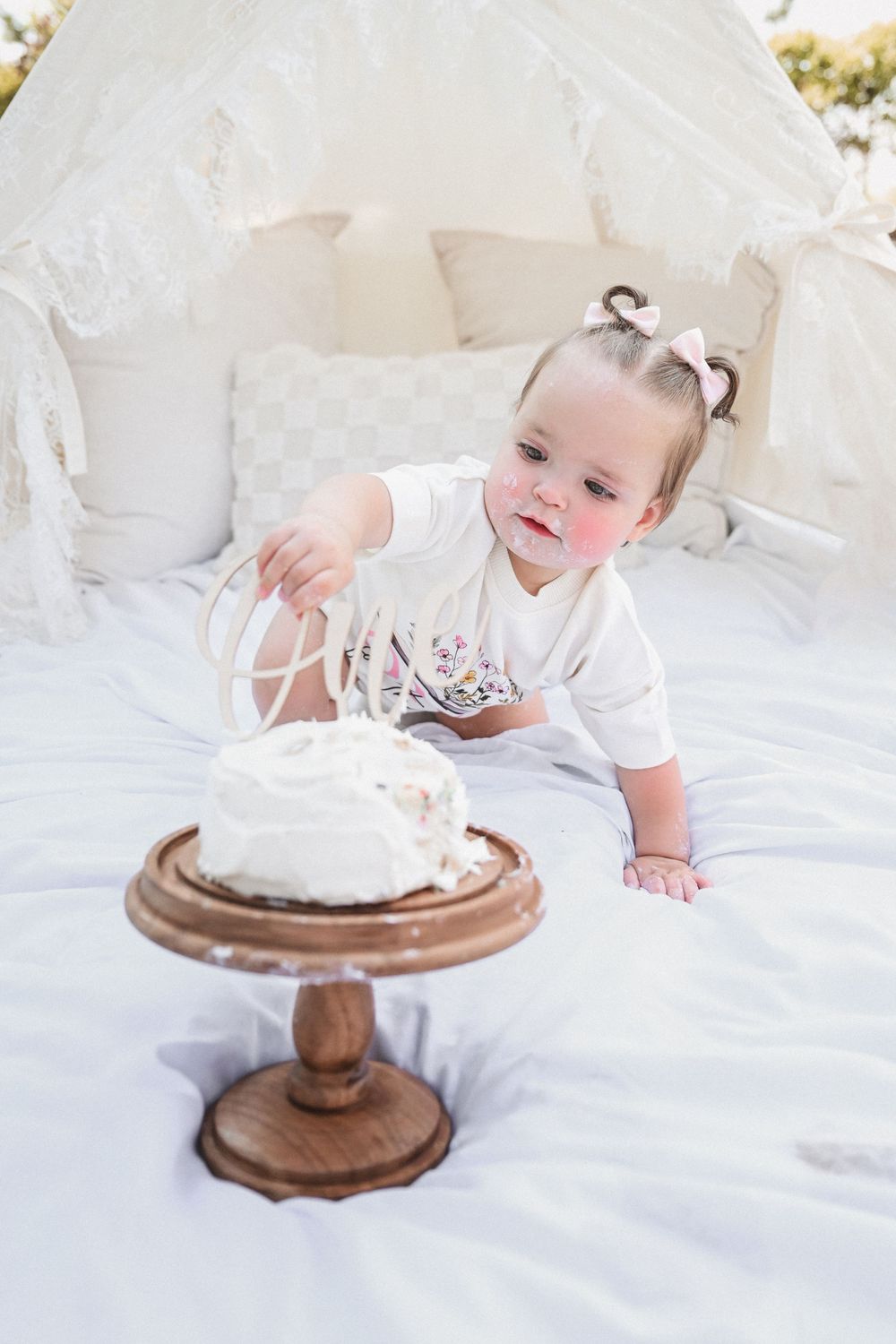 Baby in white dress reaching for cake on wooden pedestal against white backdrop.