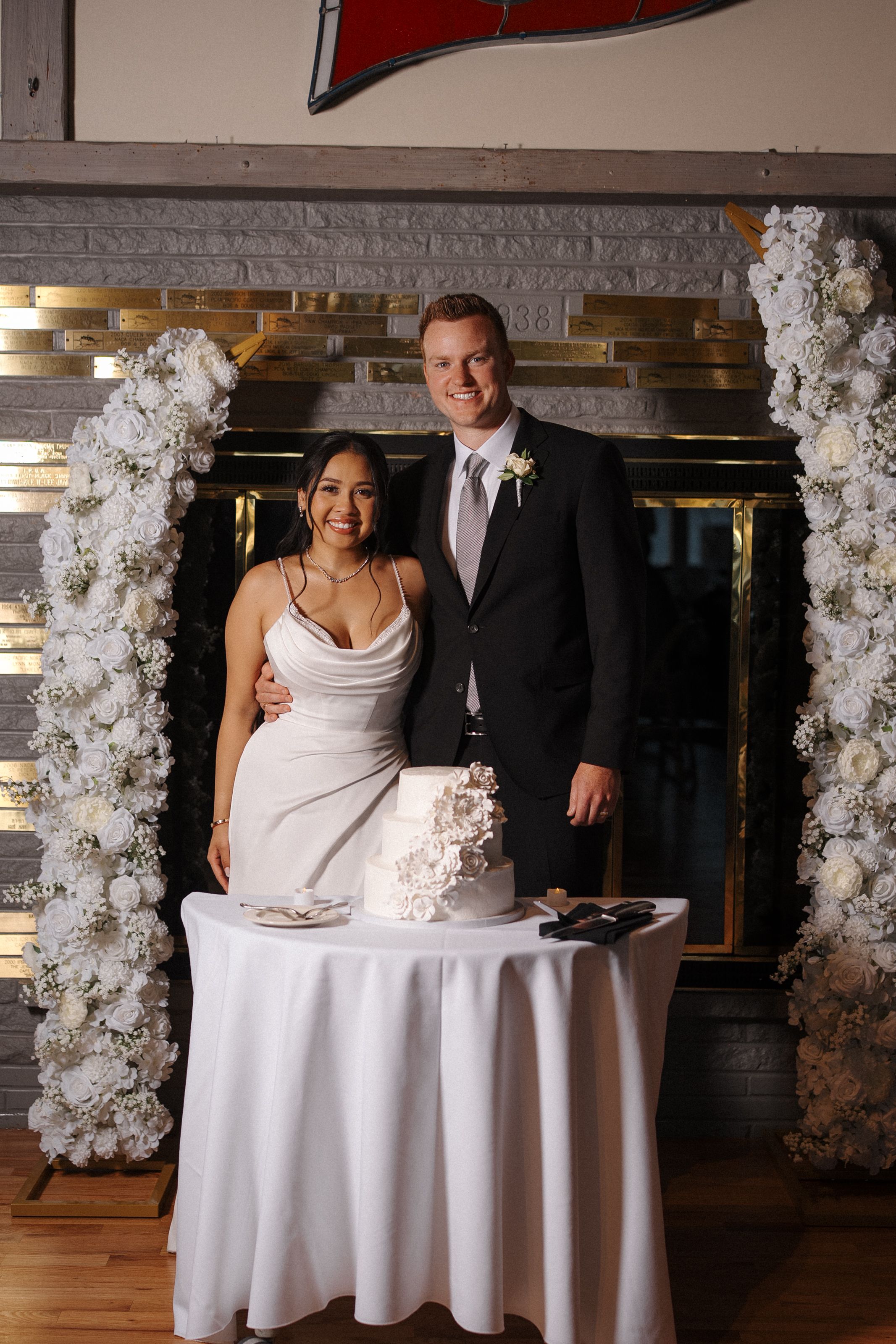 Couple stands behind round white table with wedding cake and floral decor in elegant venue with string lights.