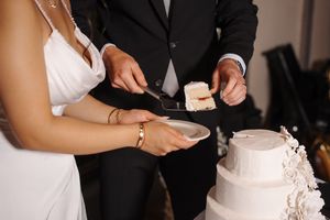 Bride and groom perform traditional cake-cutting ceremony at wedding reception, dressed in formal black and white attire.