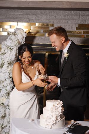 A couple shares a playful moment together while cutting their white wedding cake decorated with delicate flowers.