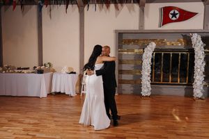 A couple dancing together in a reception hall decorated with white floral arrangements and illuminated wooden flooring.