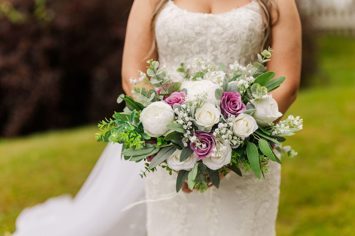 A wedding bouquet with white and purple roses, mixed greenery, and delicate flowers against a white dress and green grass.
