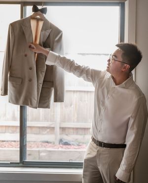 Several cream and beige colored formal suits and dress shirts hanging on display near a window.