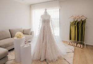 Elegant lace wedding gown displayed on mannequin in bridal boutique with bridesmaid dresses and couch in background.